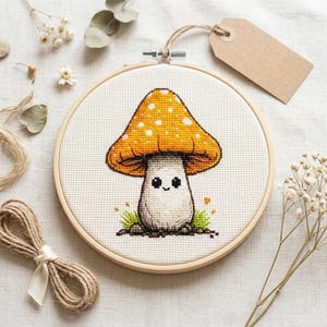 May include: A cross-stitch embroidery hoop featuring a cartoon mushroom with an orange cap and white spots. The mushroom has a smiling face and is surrounded by green grass. The embroidery is set in a wooden hoop, with a blank tag attached.