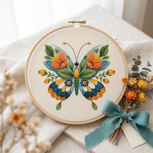 May include: A cross-stitch embroidery of a butterfly in a wooden hoop. The butterfly design features orange, blue, and green floral patterns. The embroidery is set against a cream-colored fabric. A small bouquet of dried flowers and a blue ribbon are next to the hoop.