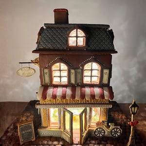 May include: A miniature gingerbread patisserie building with illuminated windows, a red and white striped awning, and a sign reading Patisserie. The structure features a grey roof, a small bicycle, a street lamp, and a sign that says OUVERT.