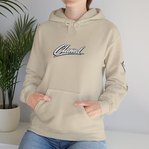 Peut inclure: Un sweat à capuche beige avec une poche avant et une capuche à cordon de serrage. Le mot "Clawed" est brodé sur la poitrine dans une police cursive. La manche a un motif graphique. Le modèle porte un jean bleu.