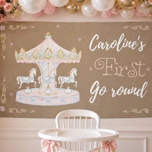 May include: A rectangular banner with a tan background featuring a watercolor illustration of a pink and blue carousel with horses. The text reads Caroline's First Go round in white script. The banner has decorative gold scrollwork corners.