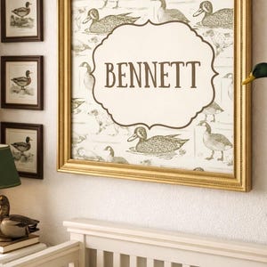 May include: A framed wall art piece with a gold frame, featuring a duck-themed pattern and the name "BENNETT" in a decorative font. The artwork is displayed above a white crib, with additional framed duck illustrations nearby.