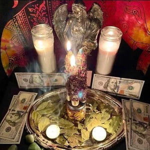 May include: A silver-toned oval tray holds a glass jar filled with dried herbs and a lit candle, surrounded by bay leaves and two small tea lights. Two tall white pillar candles, a bronze-colored angel figurine, and several 100 dollar bills are present.