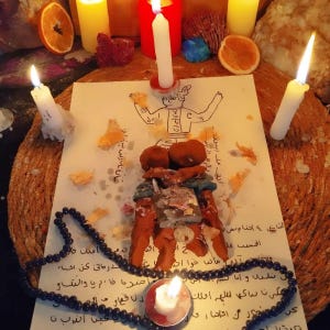 May include: A white paper with a hand-drawn figure and Arabic text, topped with a brown clay sculpture, a metal padlock, and a key. The scene is surrounded by four white candles, one red candle, two yellow candles, dried orange slices, and black prayer beads.