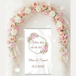 Puede incluir: Un letrero rectangular con un gráfico de corona floral con el texto "Schön dass du da bist", los nombres "Olivia & Samuel" y la fecha "12.6.2026". El letrero está enmarcado por un arco de rosas blancas y rosas y hierba de la pampa.