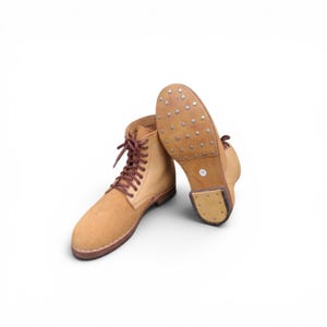 Puede incluir: Un par de botas de cuero color canela con cordones marrones. Una bota está de pie, mientras que la otra está colocada para mostrar la suela, que presenta una serie de clavos metálicos y una pequeña pegatina redonda blanca con el número 12.
