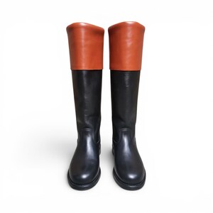 May include: A pair of knee-high boots featuring a smooth black leather lower section and a contrasting tan leather cuff at the top. The boots have a rounded toe, a flat sole, and a structured shaft that extends to the knee with visible stitching.