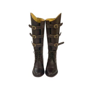 Puede incluir: Un par de botas hasta la rodilla de color marrón oscuro con cierre frontal de cordones y tres correas horizontales en la caña, cada una asegurada con una hebilla de metal dorado. El forro interior es de color beige claro y tienen punta redonda.