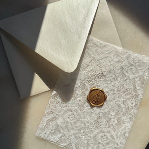 Könnte beinhalten: Ein elfenbeinfarbener Umschlag und eine weiße Spitzenkarte, versiegelt mit einem goldenen Wachssiegel mit Monogramm. Die Spitzenkarte zeigt ein detailliertes Blumenmuster. Der Umschlag ist geöffnet und gibt die Karte teilweise frei.