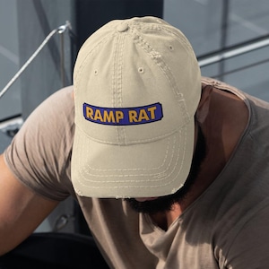 May include: A beige baseball cap with the text "RAMP RAT" in white letters on a blue rectangle. The cap is worn by a person sitting on a set of stairs. The person is wearing a brown t-shirt and dark pants.