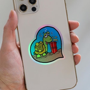 May include: A light-coloured smartphone featuring a heart-shaped sticker on its back. The sticker displays a cartoon turtle with a green shell and body, appearing dazed while standing next to a red barrel on a sandy surface against a blue background.