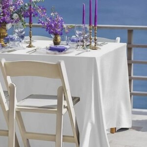 May include: A table set for a special occasion, featuring a white tablecloth, purple flowers, and candles. Gold candle holders and glassware add elegance. The scene is set against a backdrop of the sea and a wooden chair.