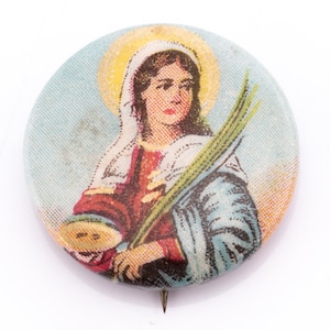 May include: A round pin with a colorful image of a woman holding a branch of leaves. The woman is wearing a blue robe and a white head covering. The background is a light blue sky.