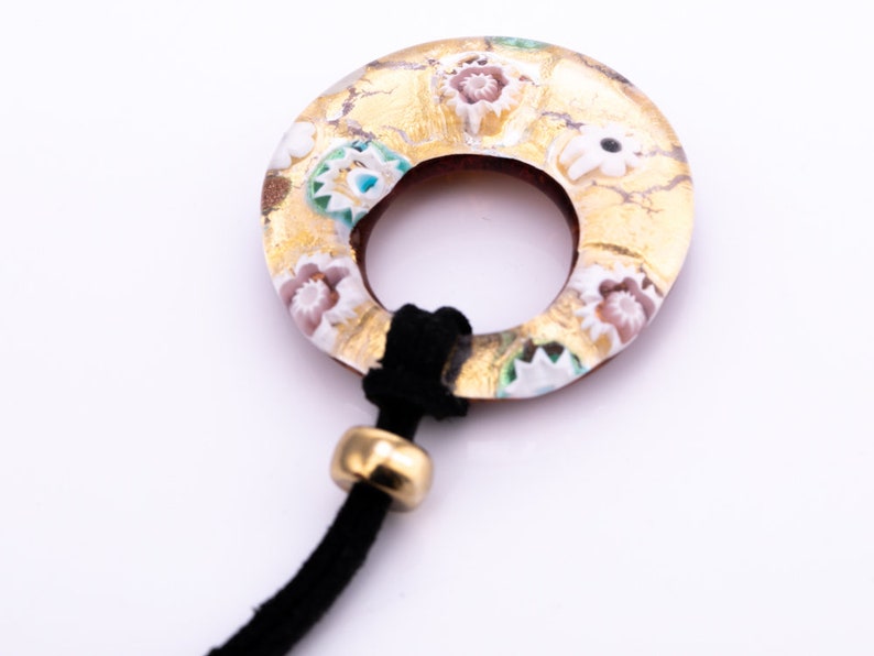 以下が含まれることがあります： 金と白のガラス製のペンダント。花柄のデザインで、黒い紐に金のビーズが付いています。