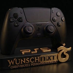 May include: A black video game controller resting on a black stand. The stand features the gold text PS5, WUNSCHTEXT, and INDIVIDUELL PERSONALISIERBAR, along with a gold logo on the right side. The controller has standard buttons and joysticks.