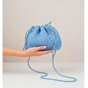 May include: A light blue drawstring pouch crafted from a textured, woven material. The bag features a gathered top secured by two thin, matching blue braided cords that extend downward, forming a long strap for carrying or hanging the item.