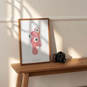 May include: A framed art print featuring a whimsical flamingo design in shades of pink and white. The artwork is displayed on a wooden bench next to a vintage black camera. The print has the text "DESIGNSYNKKVK" at the bottom.