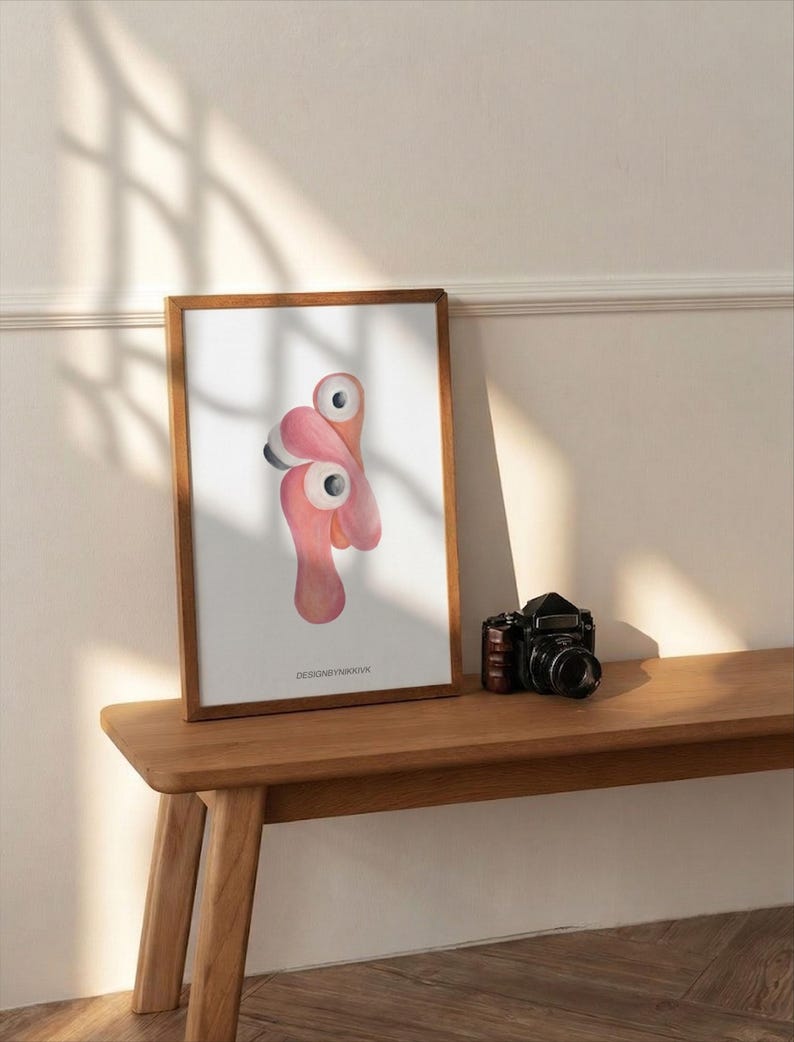 May include: A framed art print featuring a whimsical flamingo design in shades of pink and white. The artwork is displayed on a wooden bench next to a vintage black camera. The print has the text "DESIGNSYNKKVK" at the bottom.