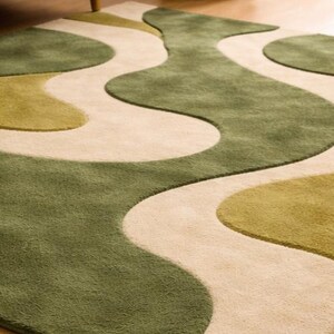 May include: A green and beige area rug with a wavy, abstract design. The rug features shades of green and beige, creating a soft, textured surface. The rug is placed on a wooden floor, with a glimpse of a sofa leg and a woven basket in the background.