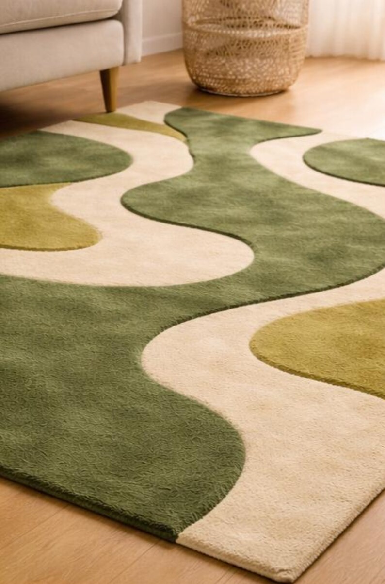 May include: A green and beige area rug with a wavy, abstract design. The rug features shades of green and beige, creating a soft, textured surface. The rug is placed on a wooden floor, with a glimpse of a sofa leg and a woven basket in the background.