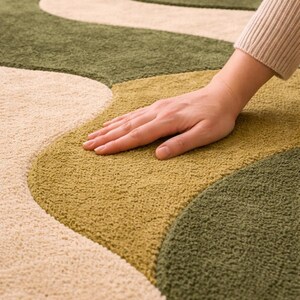 May include: A close-up of a plush, patterned rug with a wavy design. The rug features a combination of beige, olive green, and golden-yellow sections. The texture appears soft and inviting, with a hand resting on the surface.