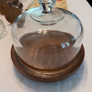 May include: A clear glass cloche with a rounded top and a clear glass handle. The cloche sits on a round, dark brown wooden base. The base has a matching dark brown wooden plate.