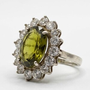 Peut inclure: Bague en argent ornée d'une grande pierre ovale vert olive, entourée d'un halo de petites pierres rondes et transparentes. La bague a un anneau en argent poli. Le design est classique et élégant.