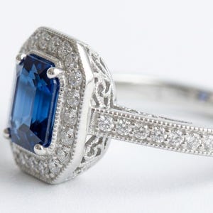 Peut inclure: Bague en argent ornée d'une pierre précieuse rectangulaire bleu foncé, entourée d'un halo de petites pierres claires. La monture présente des détails de filigrane et la bande est ornée de petites pierres.