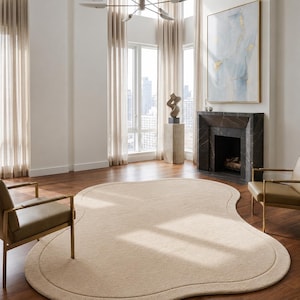 May include: A large, beige, organic-shaped area rug with a smooth, solid-coloured surface and a subtle, raised border detail that follows the irregular, flowing perimeter of the rug, positioned centrally on a dark hardwood floor in a room.