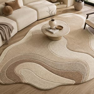 Könnte beinhalten: Beigefarbener, abstrakt geformter Teppich mit einem geschichteten Design. Der Teppich weist ein wellenförmiges Muster in Beige- und Brauntönen auf, das ein weiches, strukturiertes Aussehen erzeugt. Der Teppich liegt auf einem Holzboden und ergänzt die neutrale Farbpalette des Raumes.