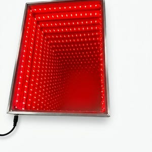 May include: A rectangular, wall-mounted light fixture with a stainless steel frame. The interior features a red, infinity mirror effect created by numerous small LED lights. A black power cord extends from the bottom left corner.