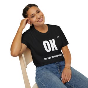 Può includere: T-shirt nera con la parola "OK" a grandi lettere bianche e la frase "ma non mi interessa" sotto. La maglietta è indossata da una persona seduta su una sedia.
