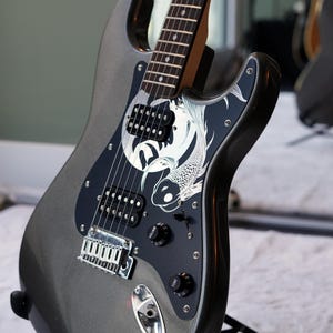 May include: A metallic grey electric guitar featuring a black pickguard adorned with a white koi fish graphic. The instrument includes two black humbucker pickups, a chrome bridge, and black volume and tone control knobs on the body.