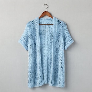May include: A light blue, short-sleeved, open-front cardigan featuring an intricate, vertical crochet knit pattern. The garment is displayed on a wooden hanger, showcasing a lightweight, semi-sheer texture with a consistent, repeating stitch design.
