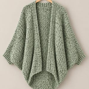 Könnte beinhalten: Eine salbeigrüne, offene Strickjacke mit einem groben, strukturierten Strickmuster. Das Kleidungsstück hat eine lockere, fallende Silhouette mit weiten Ärmeln, gerippten Bündchen und einem gerippten Kragen, präsentiert auf einem Holzkleiderbügel.