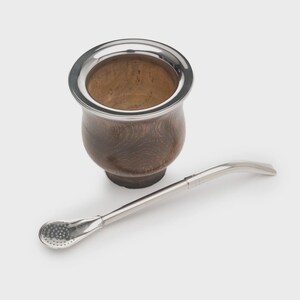 Può includere: Un contenitore per mate in legno marrone scuro con un bordo in metallo color argento lucido, abbinato a una lunga e sottile cannuccia bombilla in metallo color argento con un filtro perforato a forma di cucchiaio.