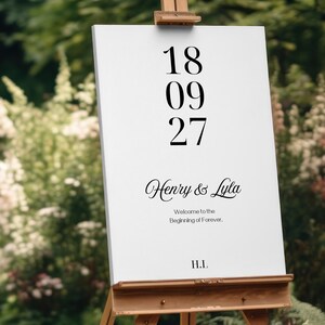 Op de afbeelding: Een wit welkomstbord voor een bruiloft op een houten ezel. Het bord toont de datum "18 09 27" in een vetgedrukte letter, de namen "Henry & Lyla" in sierlijke letters en de tekst "Welcome to the Beginning of Forever."
