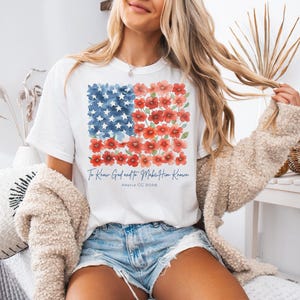 May include: A white short-sleeve t-shirt featuring a graphic of the American flag composed of blue watercolor stars and red watercolor flowers. Below the flag, the text reads To Know God and to Make Him Known and below that, Amos 6:8.