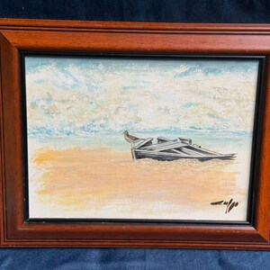 Puede incluir: Una pintura al óleo en un marco de madera marrón oscuro que representa una playa de arena con un pequeño pájaro posado sobre una estructura de barco de madera desgastada y parcialmente sumergida. El cielo tiene tonos azul claro y blanco.