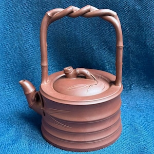 Puede incluir: Una tetera de arcilla marrón con cuerpo cilíndrico, ranuras horizontales y detalles inspirados en el bambú. La tetera incluye un asa alta y arqueada con diseño trenzado, un pico corto y una tapa adornada con un pequeño motivo de hoja.