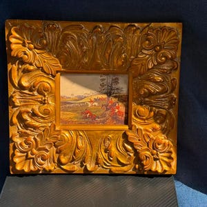 Puede incluir: Una pintura rectangular que muestra una escena de caza del zorro con jinetes a caballo en un campo, encerrada en un marco grueso, ornamentado y de color marrón dorado con tallas en relieve de hojas de acanto y patrones de volutas.