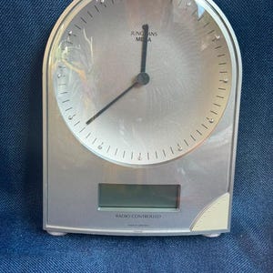 May include: A silver-toned Junghans Mega radio-controlled table clock with an arched top. The clock face features a metallic finish, thin black hands, and small markers. Below the dial is a rectangular digital display and the text "RADIO CONTROLLED".