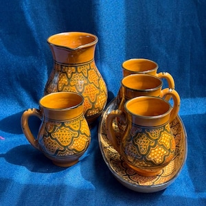 Puede incluir: Un juego de bebidas de cerámica que consta de una jarra, tres tazas y una bandeja ovalada. Cada pieza presenta un color base naranja cálido con un patrón festoneado repetitivo en verde oscuro y amarillo. La jarra tiene un pico ancho.