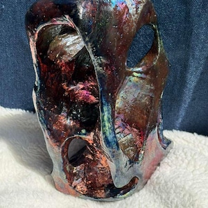 Puede incluir: Una escultura de cerámica artesanal con una forma orgánica única. La pieza presenta una mezcla de colores, incluyendo rojos profundos, morados y azules, con reflejos iridiscentes. Tiene múltiples aberturas y una superficie texturizada.