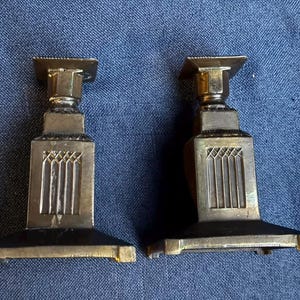Puede incluir: Un par de candelabros de latón a juego con base cuadrada, cuerpo cónico con un patrón de celosía vertical en cada lado y una plataforma superior cuadrada. El metal tiene un acabado pulido con signos visibles de antigüedad y pátina.
