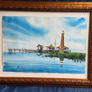 May include: Watercolour painting of a coastal scene with a lighthouse, pier, and small buildings. The artwork is framed in a gold-toned wooden frame. The sky is blue with white clouds, and the water reflects the scene.