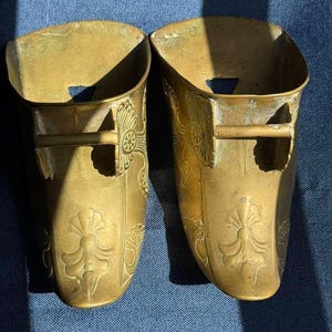 May include: A pair of matching brass stirrups featuring an open-top design with a horizontal bar across the front. The exterior surface of each stirrup is decorated with embossed floral and fan-shaped patterns, showing a metallic gold finish.