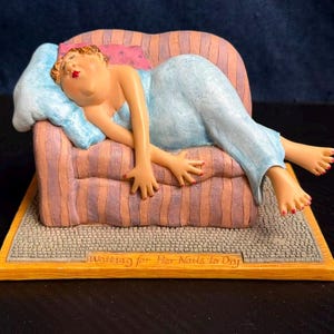 May include: A figurine of a person in a light blue dress resting on a striped pink and purple armchair. The person has red nail polish and is lying on a light blue pillow. The base features a textured gray surface with the text Waiting for Her Nails to Dry.
