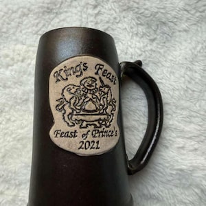 Puede incluir: Una jarra de cerveza de cerámica marrón oscuro con asa. La jarra presenta un emblema de color crema con las palabras "King's Feast" y "Feast of Prince's 2021" en letras negras, junto con una ilustración decorativa.
