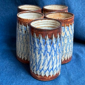 Puede incluir: Un juego de cinco vasos cilíndricos de cerámica con un patrón de chevrón azul y blanco texturizado y repetitivo. Los bordes y las bases tienen un acabado de esmalte marrón rojizo terroso, con una superficie interior de color blanco roto moteado.
